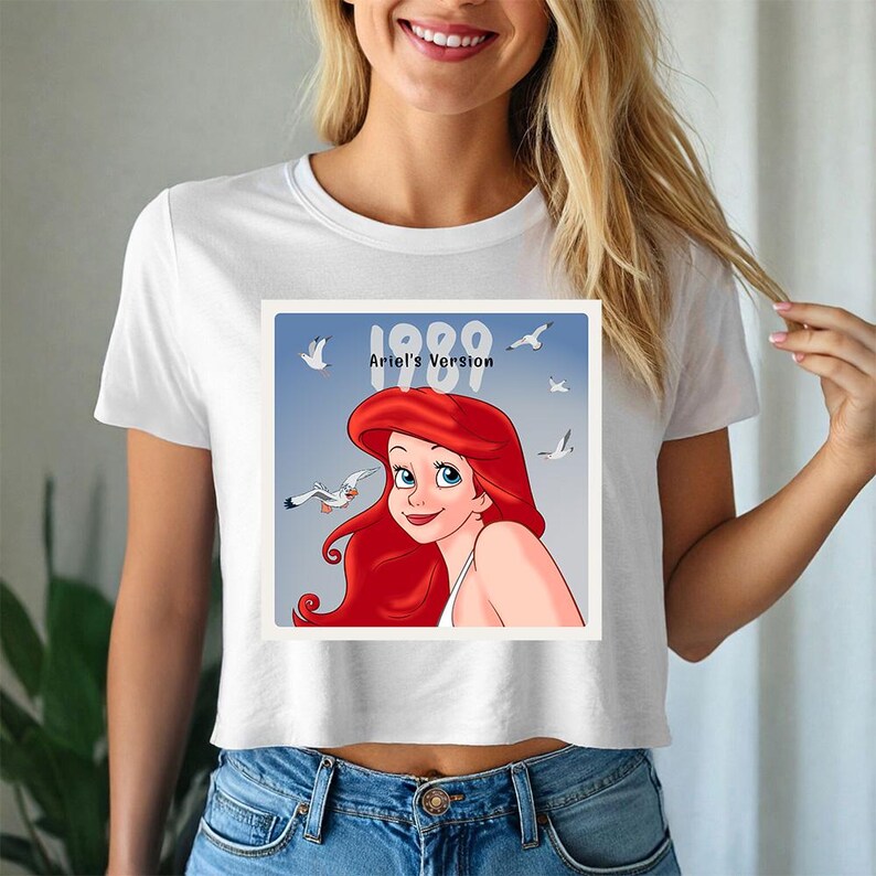 May include: White crop top t-shirt featuring a graphic of Ariel from The Little Mermaid. The graphic includes the text "1989 Ariel's Version" with Ariel's image and seagulls on a blue background. The shirt is paired with blue jeans.