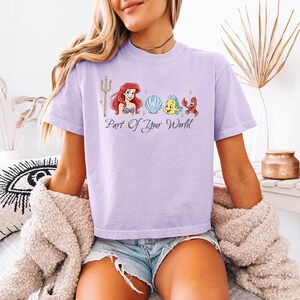 May include: A light purple t-shirt featuring a graphic of Ariel, Flounder, Sebastian, and a seashell. The phrase "Part of Your World" is printed below the characters. The shirt is a short-sleeved, crew neck style.