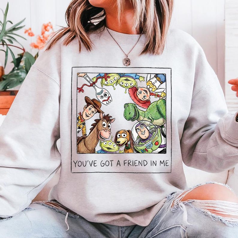 May include: A pale grey sweatshirt with a colourful illustration of characters from the animated film "Toy Story." The image includes Woody, Buzz Lightyear, Rex, and other characters, with the text "YOU'VE GOT A FRIEND IN ME" below the image.