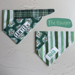 May include: Two triangular pet bandanas featuring green and white striped fabric and a green patterned fabric with squirrel illustrations. One bandana has a white patch with the name Sawyer, and the other has a white patch with the name lola.