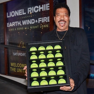 May include: A black framed display case filled with 25 bright yellow tennis balls. The case is being held by a man wearing a black jacket and a gold chain.