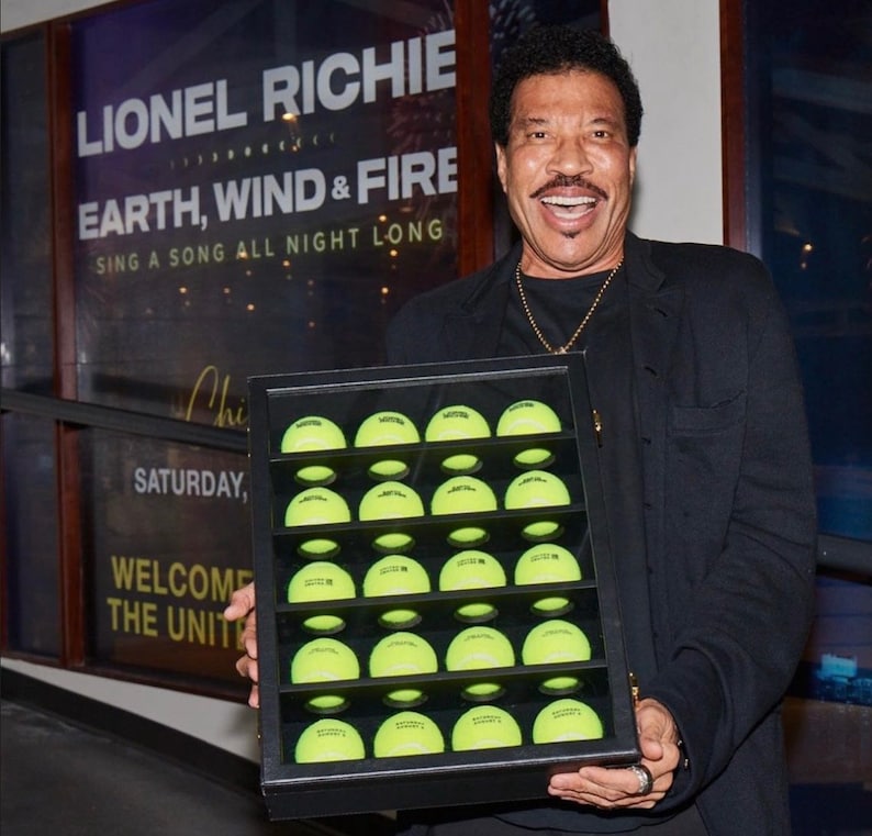 May include: A black framed display case filled with 25 bright yellow tennis balls. The case is being held by a man wearing a black jacket and a gold chain.