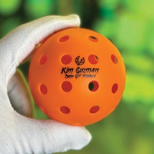 May include: An orange pickleball with multiple circular holes. The ball has the text "Kim Guzman Come Get Smoked" printed on it in black. The ball is held in a white gloved hand against a blurred green background.