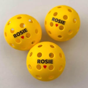 May include: Three yellow pickleballs with multiple circular holes. Each ball has the name "ROSIE" printed in black, with a small red heart below the name. The balls are arranged on a light-colored surface.