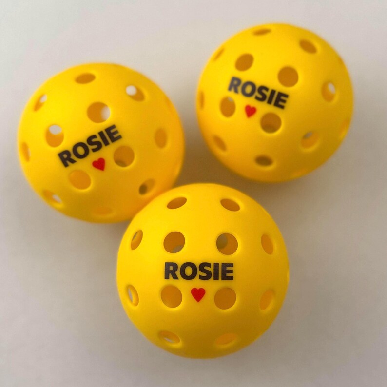 May include: Three yellow pickleballs with multiple circular holes. Each ball has the name "ROSIE" printed in black, with a small red heart below the name. The balls are arranged on a light-colored surface.