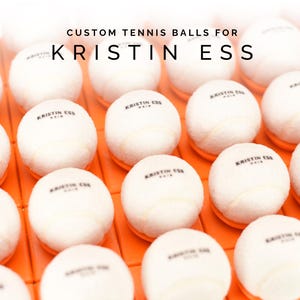 May include: A collection of white tennis balls with black lettering that reads "KRISTIN ESS HAIR" on each ball.