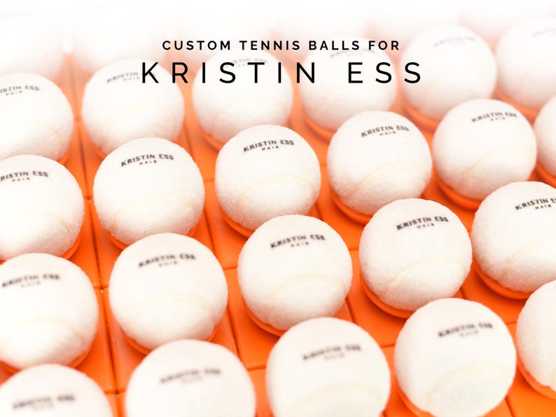 May include: A collection of white tennis balls with black lettering that reads "KRISTIN ESS HAIR" on each ball.
