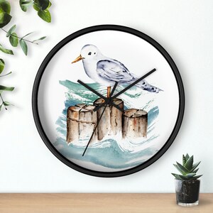 May include: A black framed clock with a white background featuring a watercolour painting of a seagull perched on wooden posts in the ocean. The clock hands are black.
