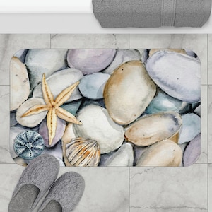 May include: A watercolor bath mat with a beach theme. The mat features a variety of smooth, round stones in shades of gray, blue, and brown. There is a starfish, a seashell, and a sea urchin on the mat.