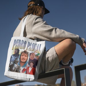 Peut inclure: Un sac fourre-tout blanc avec un imprimé graphique coloré comportant le texte "PEDRO PASCAL" et des images d'un homme avec une moustache et une barbe, d'un homme avec une barbe et une moustache et d'un homme avec une barbe et une moustache. Le sac a une bandoulière blanche.