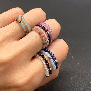 May include: A hand displays a collection of beaded rings in various colors. The rings feature small, colorful beads strung together, creating a delicate and stylish accessory. Colors include clear, black, pink, purple, blue, and green.