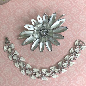 May include: A silver-toned metal flower brooch and a bracelet with white enamel leaf accents. The jewelry is displayed on a pink floral patterned surface. The brooch has a central crystal-like embellishment.