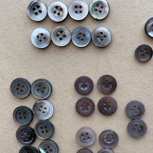 May include: A collection of iridescent mother-of-pearl buttons in various sizes. The buttons have four holes and a subtle, pearlescent sheen. The arrangement includes several sets of buttons, showcasing their different sizes and colors.