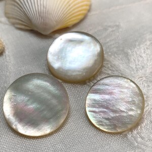 May include: Three iridescent, round mother-of-pearl buttons with a pearlescent sheen. The buttons are arranged on a white, textured surface, with a seashell in the background. The buttons have a natural, organic look.