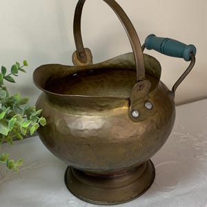 May include: Antique brass bucket with a hammered texture, featuring a curved handle and a teal-colored wooden handle. The bucket has a rounded body and a pedestal base, showcasing a vintage aesthetic.