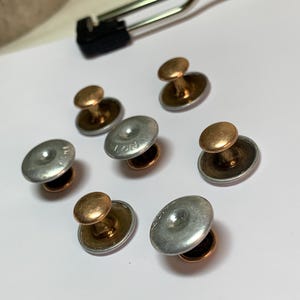 May include: A collection of seven vintage shirt studs. Each stud features a silver-colored, flat, circular top with a smaller, gold-colored, rounded top. The studs are arranged on a white surface, with a metal clip in the background.