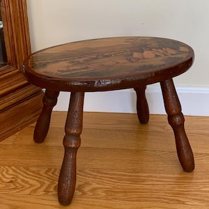 May include: A small, oval-topped wooden stool with a dark brown finish. The top features a landscape painting. The stool has three turned legs. The wood grain is visible throughout the piece, giving it a rustic appearance.