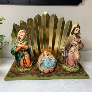 May include: A nativity scene featuring three painted figurines on a green base. A figure in a green and orange robe stands on the left, a figure in a brown robe holds a lantern on the right, and a baby in a blue cloth lies in the center. A gold fan-shaped backdrop stands behind the figures.