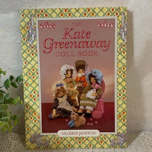 May include: The Kate Greenaway Doll Book by Valerie Janitch. The cover features illustrations of dolls in period clothing. The book has a pink background with the title in red and white lettering. The border is yellow with a repeating pattern.