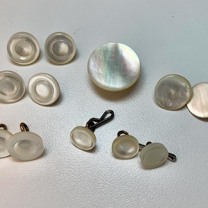 May include: A collection of vintage mother-of-pearl cufflinks and buttons. The cufflinks have black metal clasps, and the buttons are round and oval. The iridescent surfaces reflect light, showcasing a range of colors.