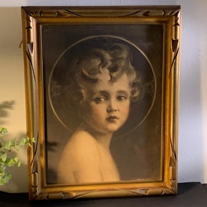 May include: A framed portrait of a young child with curly hair and a halo, rendered in soft sepia tones. The artwork is enclosed in a rectangular, ornate gold-colored wooden frame with carved geometric details along the edges.
