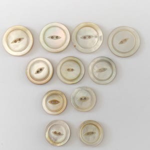 May include: A collection of round, iridescent mother-of-pearl buttons. Each button has two small holes for sewing. The buttons vary slightly in color, ranging from white to cream, and are arranged on a white background.
