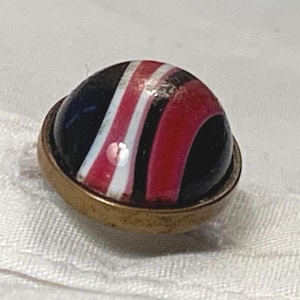 May include: A vintage marble with a red, white, and blue striped pattern, encased in a brass-colored metal setting. The marble has a glossy finish and is set against a white fabric background. The design is reminiscent of a patriotic theme.