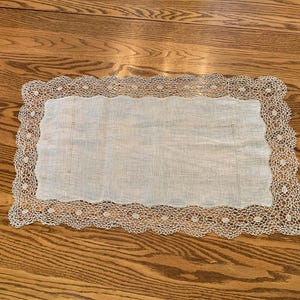 May include: A rectangular, off-white lace doily with a scalloped edge. The center is a plain, light-colored fabric. The doily is placed on a wooden surface, showcasing its delicate design and texture.