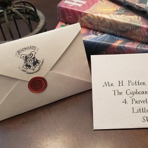 Può includere: Una lettera di accettazione di Hogwarts color crema con un sigillo di cera rosso e una carta separata con un indirizzo scritto a mano. La lettera è accanto a una bacchetta e libri.