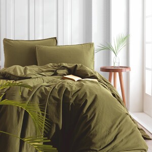 May include: A green duvet cover with a book on top of it. The duvet cover is on a bed with two pillows. There is a plant in the foreground and a small table with a vase of greenery in the background.