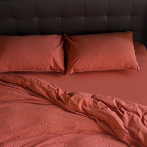 May include: A rust-colored bed with a dark brown headboard and two matching rust-colored pillows. The bed is made with a rust-colored duvet cover and the duvet is slightly pulled back to reveal the fitted sheet.