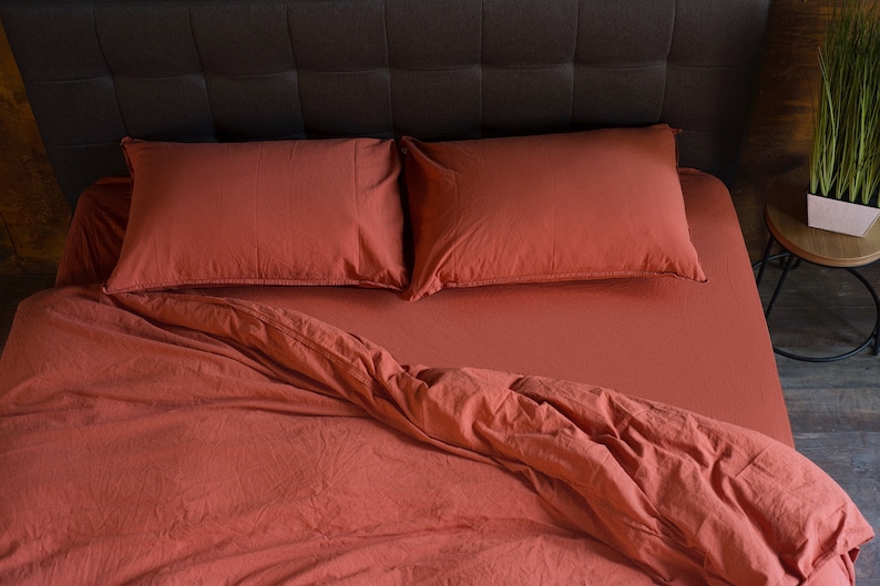May include: A rust-colored bed with a dark brown headboard and two matching rust-colored pillows. The bed is made with a rust-colored duvet cover and the duvet is slightly pulled back to reveal the fitted sheet.