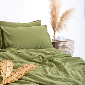 May include: A green duvet cover and pillowcases with a pair of dried palm leaves on top. The bedding is on a bed with a white wall in the background. There is a wicker basket with dried pampas grass next to the bed.