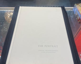 May include: A hardcover book titled "THE PORTRAIT" with a white cover. The book's foreword is by Nelson Mandela, and the author is Rosalind Coward. The book rests on a black, textured surface.