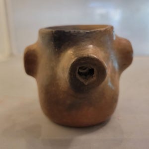 May include: A small, handmade, brown ceramic vessel with a rounded body and two side handles. The vessel has a dark, circular opening on the front and a dark interior. The ceramic piece has a rustic, earthy aesthetic.