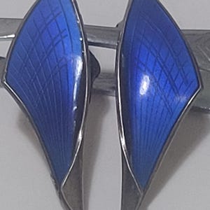 May include: A pair of vintage earrings with a silver-tone metal frame and vibrant blue enamel inlay. The enamel has a textured, grid-like pattern. The earrings have a unique, wing-like shape, adding a touch of elegance.