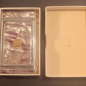 May include: A vintage cigarette case with a silver-tone frame and a pearlescent purple and white interior. The case features a gold-colored emblem and is presented in its original off-white box.