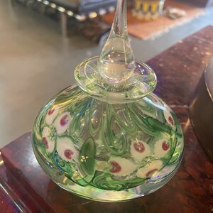 May include: A clear glass bottle with a green and white floral design. The bottle has a glass stopper with a teardrop shape.