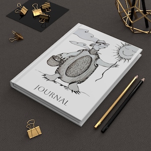 May include: White journal with the word "JOURNAL" printed on the cover. The cover features a whimsical illustration of a turtle carrying a basket and a smiling sun. Gold and black pencils and gold binder clips are also visible.