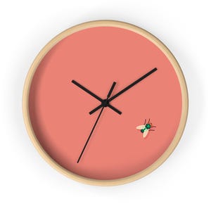 May include: Round wall clock with a salmon-colored face and a light wood frame. The clock features black hands and a small illustration of a fly. A decorative item for any room.