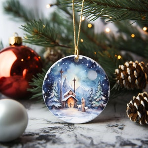 K&ouml;nnte beinhalten: Ein wei&szlig;er Keramik-Weihnachtsbaumschmuck mit einem Aquarellbild einer verschneiten Berglandschaft. Der Anh&auml;nger zeigt eine kleine Kirche mit einem Stern oben, einen Hirten mit einem Stab und zwei Schafe. Der Hintergrund ist ein dunkelblauer Himmel mit wei&szlig;en Sternen und einem Vollmond.