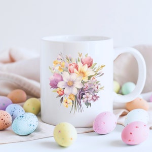 May include: White ceramic mug with a floral design featuring pink, yellow, and purple flowers. The mug is surrounded by pastel-colored Easter eggs.