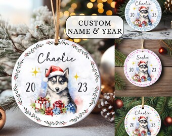 May include: A white ceramic Christmas ornament with a watercolor illustration of a husky dog wearing a Santa hat. The ornament has a white background with a green wreath around the edge. The dog is surrounded by presents and the text "Charlie" is written at the top of the ornament. The years "20" and "23" are written on either side of the dog.