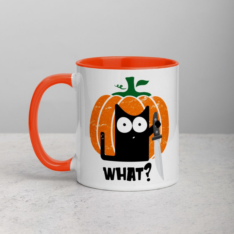 May include: White ceramic mug with an orange rim. The mug features a black cat holding a knife, peeking out from behind a cartoon pumpkin. The text "WHAT?" is printed below the cat.