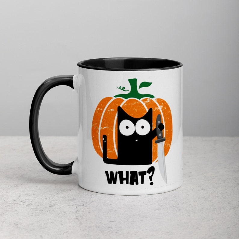 May include: White ceramic coffee mug with a black rim and handle. The mug features a black cat holding a knife, peeking out from behind a cartoon pumpkin with the text "WHAT?" printed below.