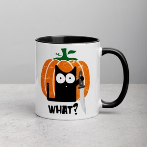 May include: White ceramic mug with a black rim and handle. The mug features a black cat holding a knife, peeking out from behind a cartoon orange pumpkin with a green stem. The text "WHAT?" is printed below the cat.