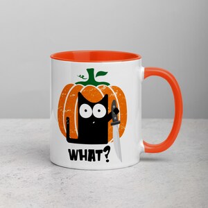 May include: A white ceramic mug with an orange rim and handle. The mug features a black cat with a knife inside a pumpkin with the text "WHAT?" printed on the front.