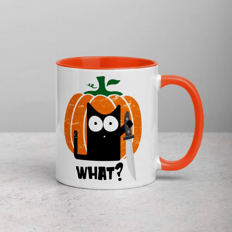 May include: A white ceramic mug with an orange rim and handle. The mug features a black cat with a knife inside a pumpkin with the text "WHAT?" printed on the front.