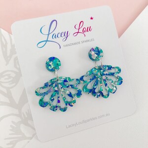 May include: Teal and blue glitter dangle earrings. The earrings feature a circular stud and a fan-shaped design with cut-out details. The earrings are displayed on a white card with the text "Lacey Lou Handmade Sparkles".