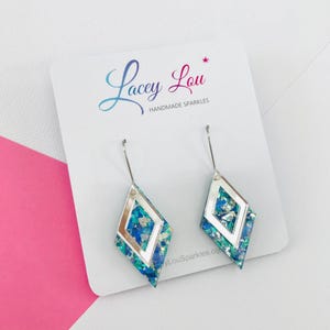 May include: Diamond-shaped earrings with a silver frame and a blue and green glitter design. The earrings are displayed on a white card with the text "Lacey Lou Handmade Sparkles".